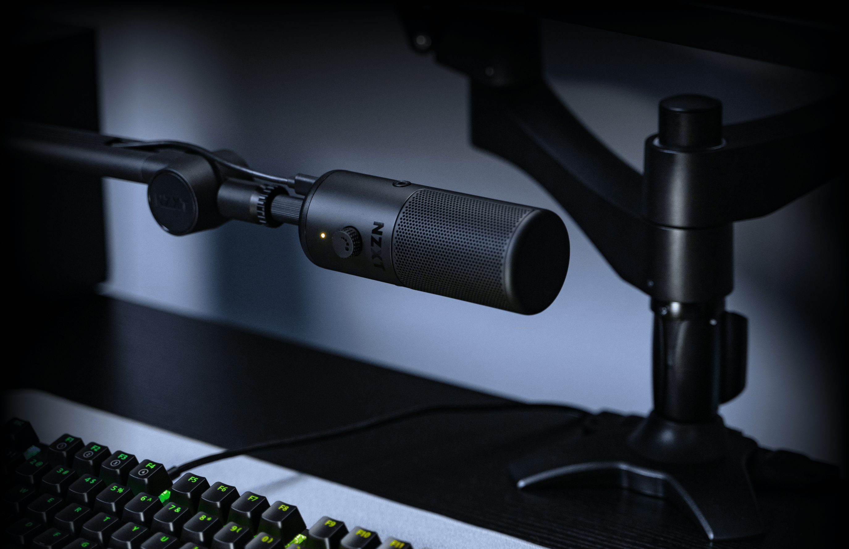 A black condenser microphone on an adjustable arm stand is positioned above a black computer keyboard on a dark desk, with part of a monitor mount visible in the background.
