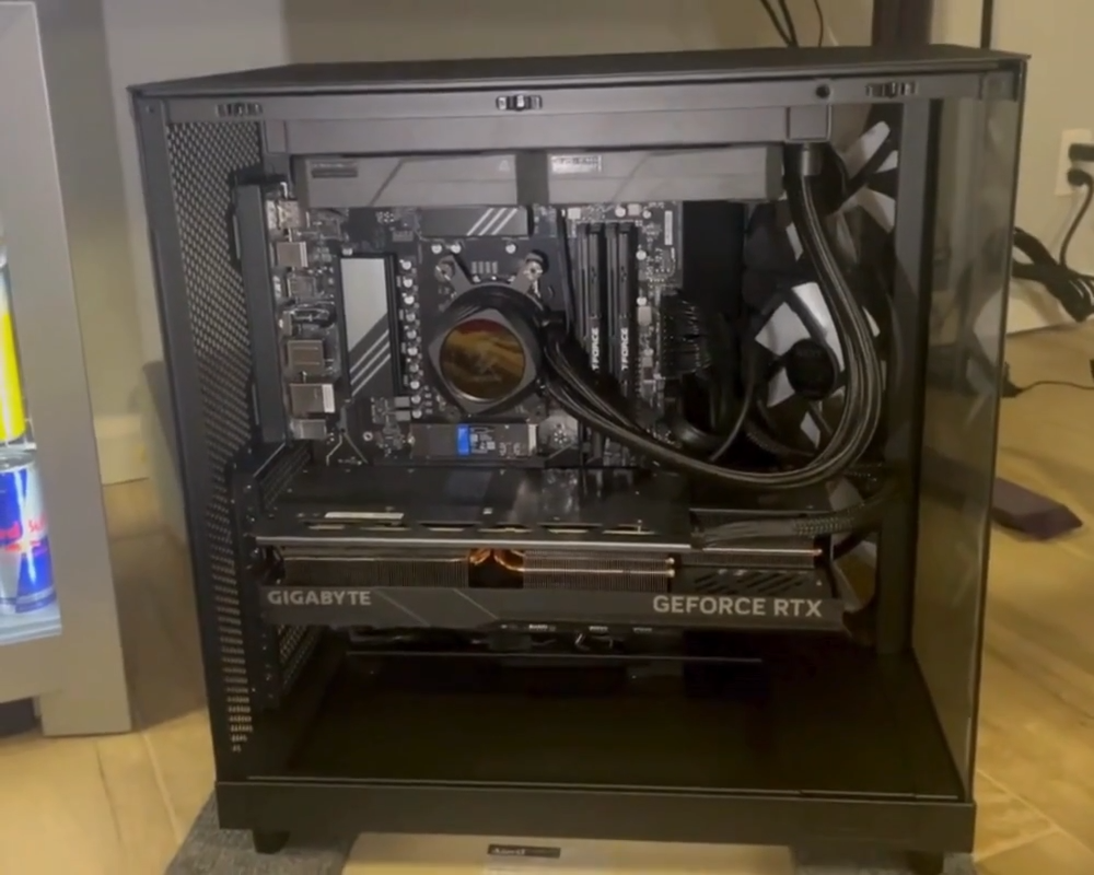 A black desktop gaming PC with a transparent side panel reveals internal components, including a GeForce RTX graphics card, water cooling system, and motherboard. The case is placed on a light wooden floor near a mini fridge.