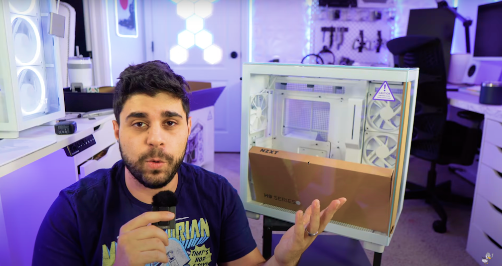 A man holding a microphone sits in front of a white computer case with an NZXT H9 Series box inside. He gestures with one hand, and the room is filled with tech equipment and purple lighting.