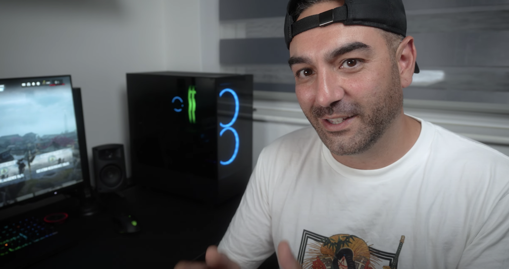 A man wearing a white T-shirt and backwards cap sits at a desk with a gaming PC and monitor displaying a video game in the background. He is smiling and gesturing towards the camera.
