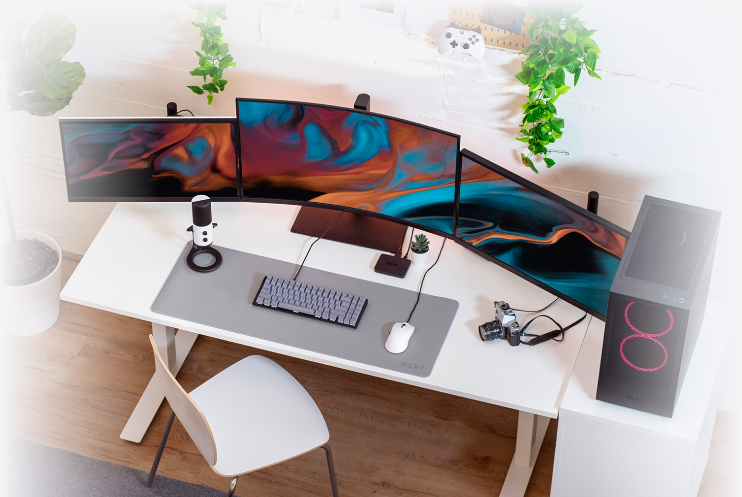 A modern desk setup with three curved monitors, a mechanical keyboard, mouse on a grey mat, microphone, camera, plant, and a gaming PC with red lights, in a bright room with hanging plants.
