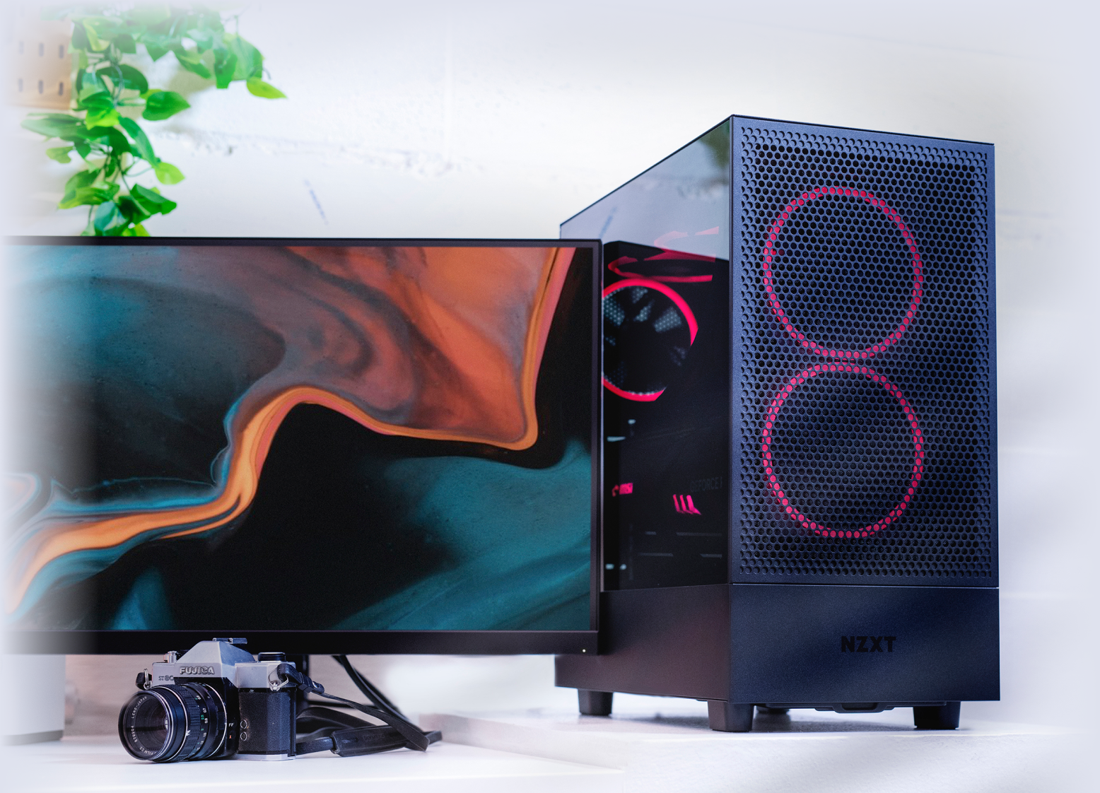 A modern desktop computer with red LED lights in the case sits beside a widescreen monitor displaying abstract art. A vintage camera and a plant are also on the white desk.