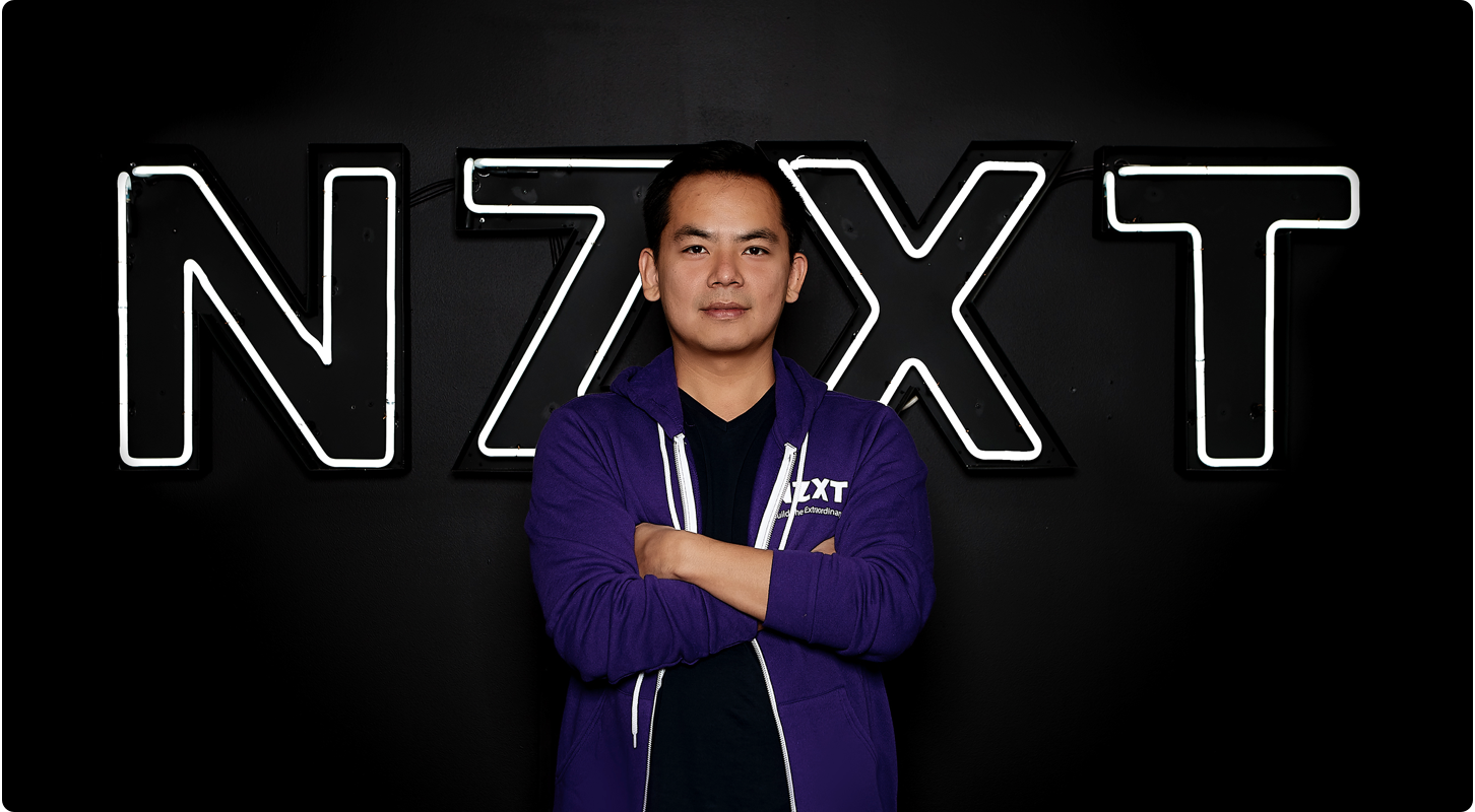 A person wearing a purple NZXT hoodie stands with arms crossed in front of a large illuminated NZXT sign on a black background.