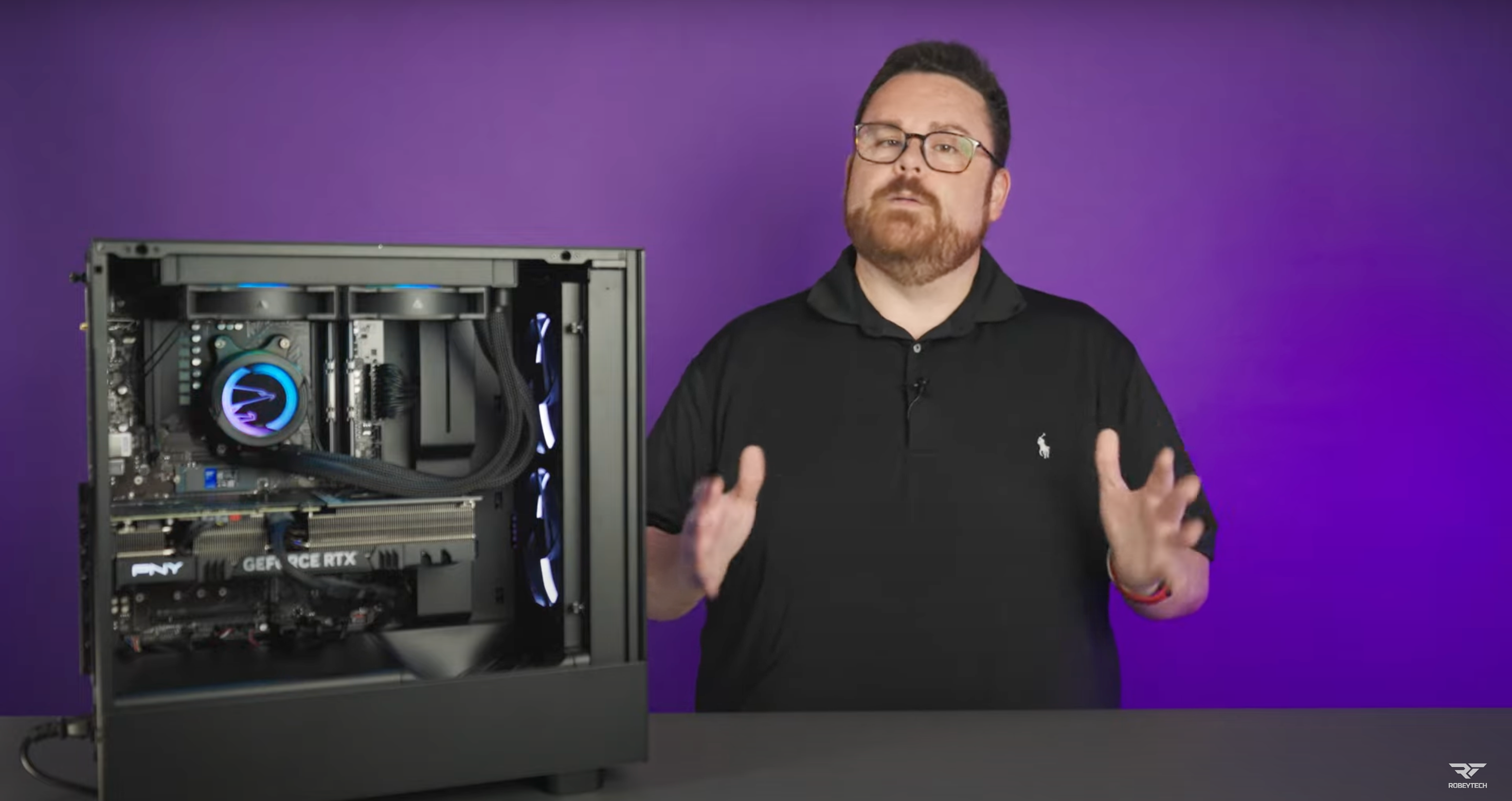 A man with glasses and a beard stands next to an open desktop computer, gesturing with his hands. The computer’s internal components are visible. The background is a solid purple color.