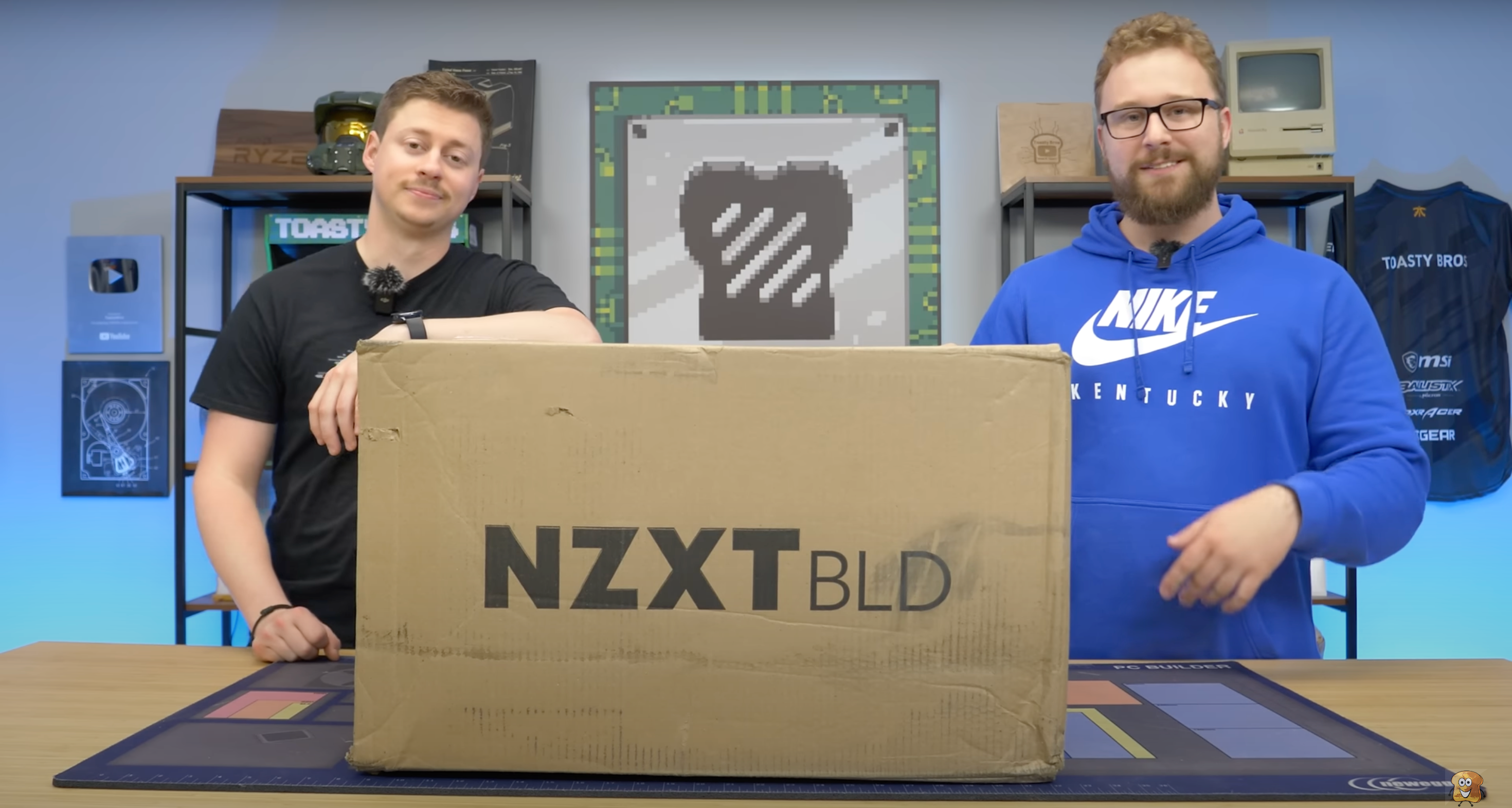 Two men stand behind a large cardboard box labeled NZXT BLD on a table, with tech-themed decor and computer parts visible in the background.